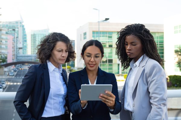 Concentrated businesswomen with digital tablet. Focused multiethnic female colleagues standing and using tablet pc together on street. Technology concept
